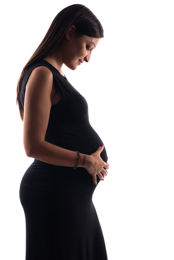 Foto ritratto controluce di profilo di una donna in gravidanza con vestito nero attillato che si tocca la pancia. sfondo bianco