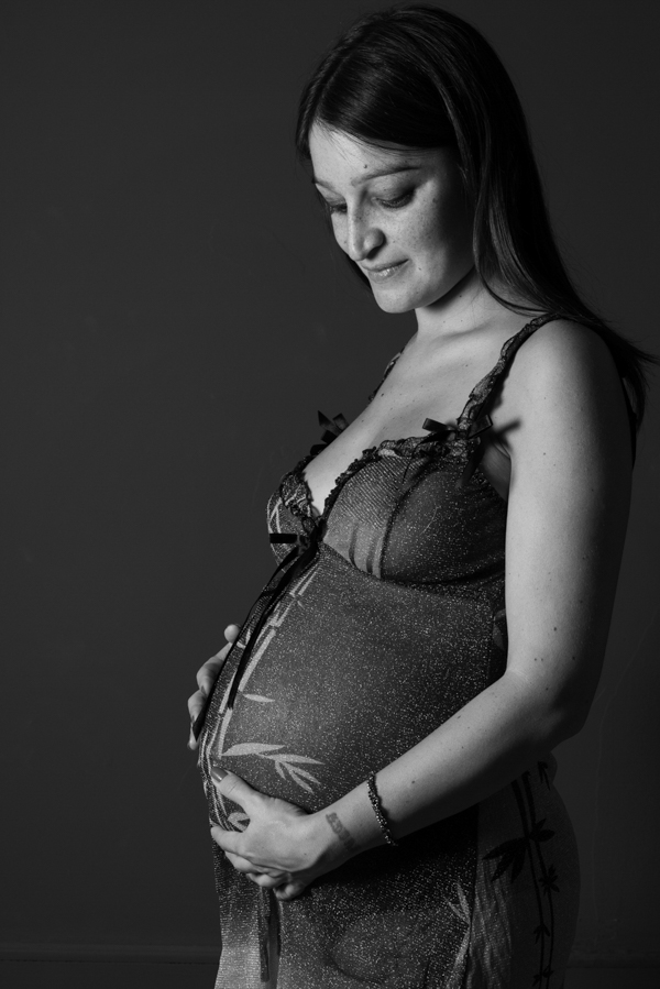 Ritratto in bianco e nero su sfondo scuro di una donna in gravidanza con baby doll che si guarda la pancia sorridendo
