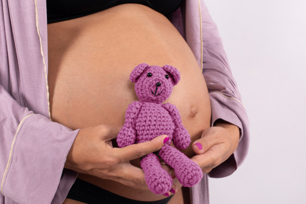 Dettaglio della pancia di una mamma in dolce attesa, con le mani che tengono un pupazzetto rosa fatto a uncinetto