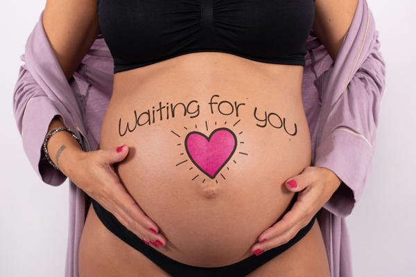 Primo piano frontale della pancia di una donna in gravidanza con scritto sopra waiting for you e un cuoricino rosa