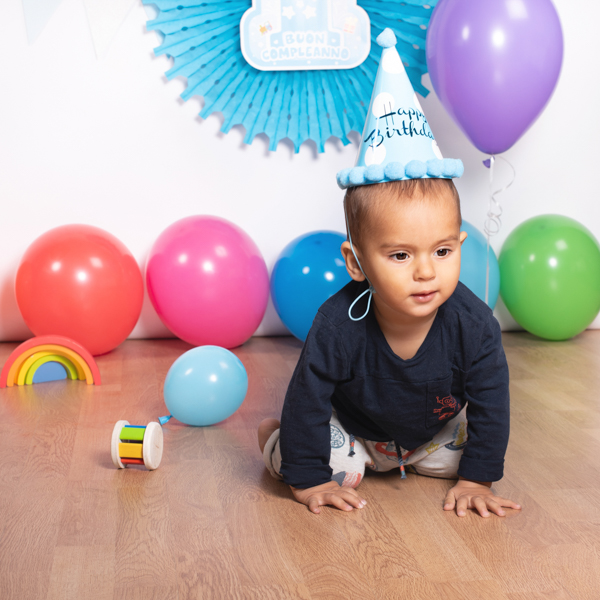 foto di un bimbo al suo primo compleanno, con cappellino da festa e palloncini colorati