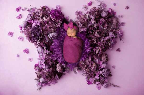 Foto artistica di neonata avvolta in una stoffa rosa scuro, con fiocco in testa, al centro di una composizione di fiori viola a forma di farfalla