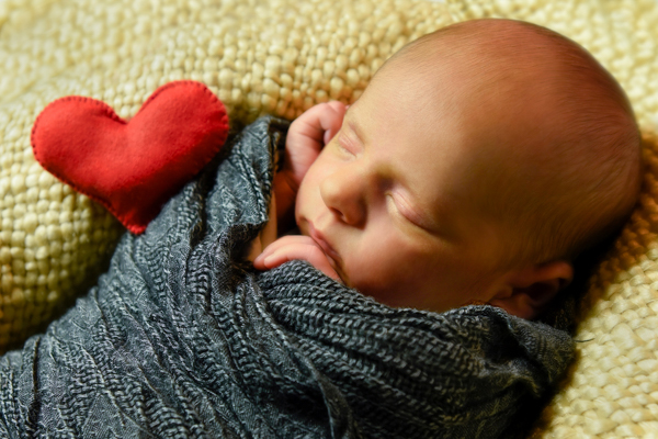 Foto newborn con primo piano del bambino avvolto in stoffa grigio scuro, su copertina di lana gialla e cuoricino di stoffa rosso