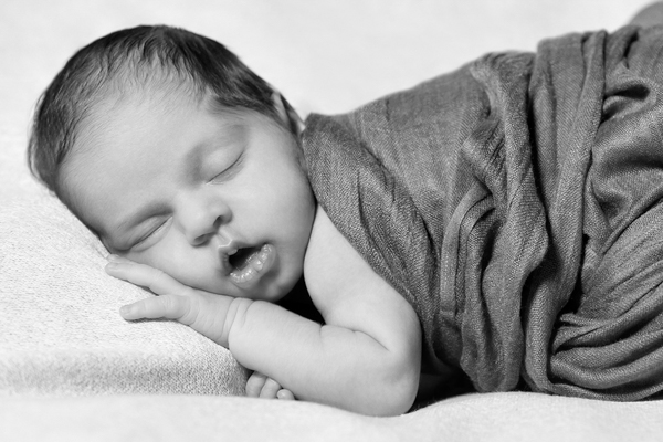 Primo piano di una neonata a pancia in giù, che dorme col viso appoggiato su una manina, avvolta in stoffa morbida. Bianco e nero