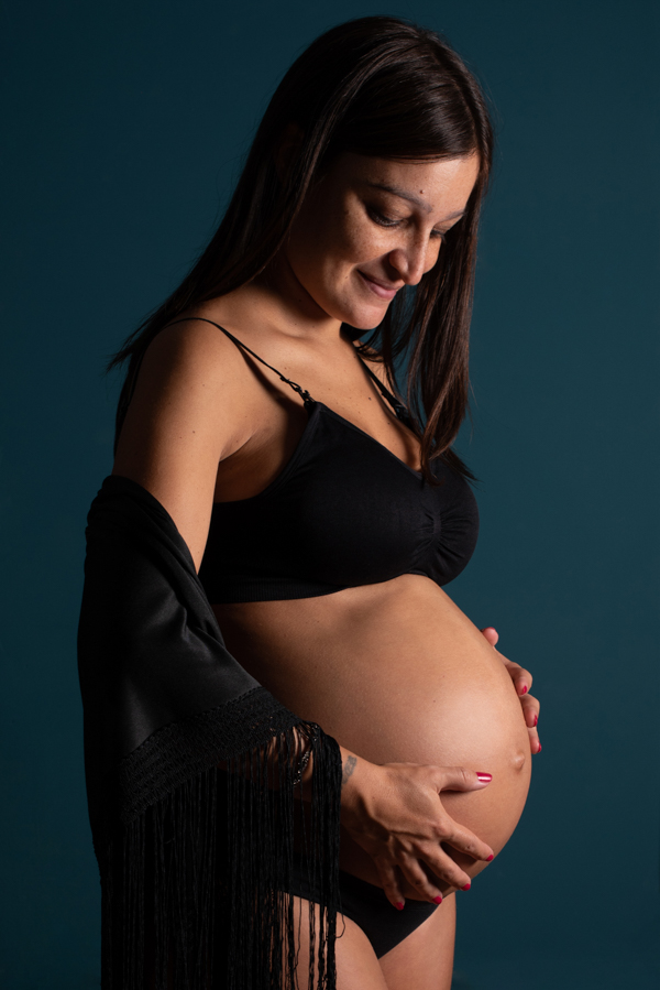 Foto ritratto di una futura mamma che si tocca il pancione sorridendo. Sfondo verde scuro e luce laterale