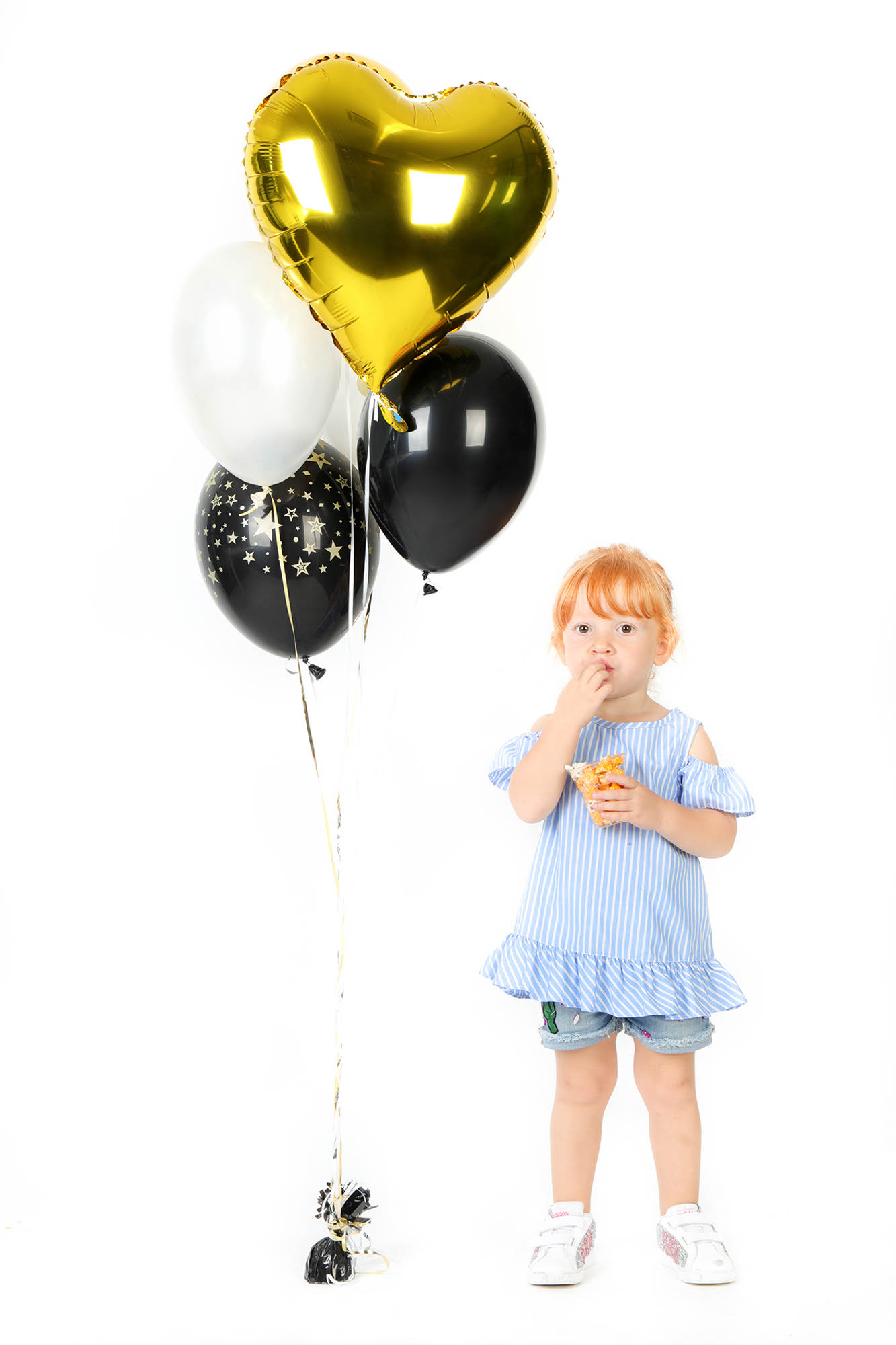 foto di una bimba rossa che mangia, in piedi, accanto a 4 palloncini