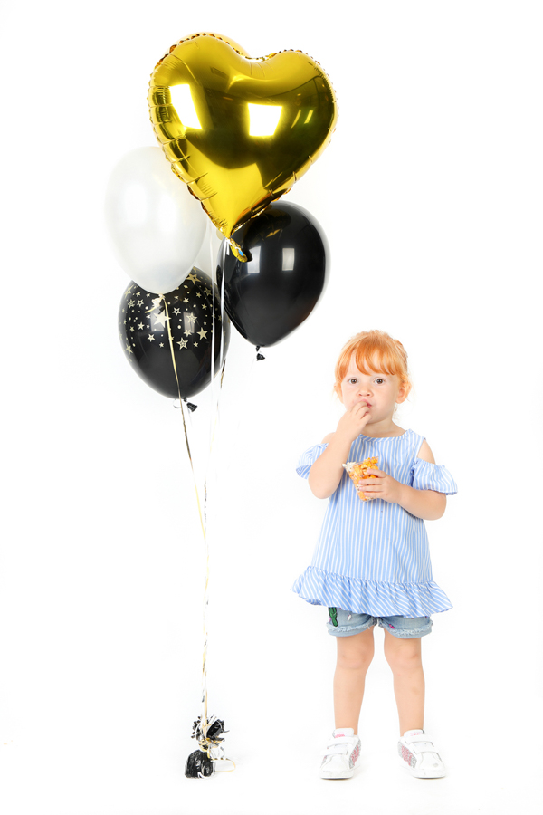 foto di una bimba rossa che mangia, in piedi, accanto a 4 palloncini