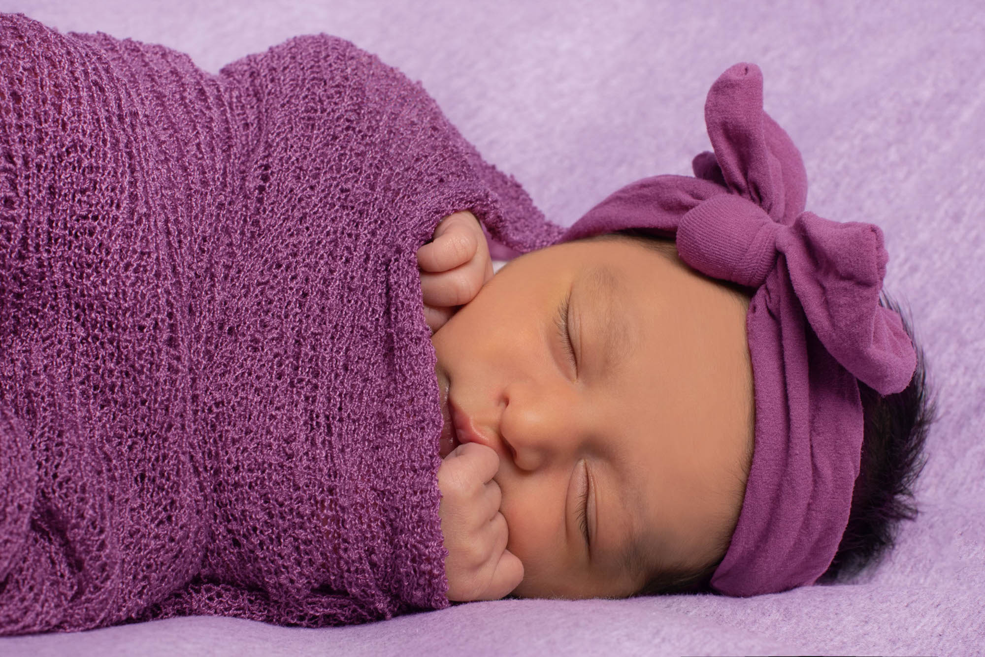 Primo piano neonata che dorme, avvolta in stoffa viola, con le manine che sbucano, e fiocco in testa dello stesso colore