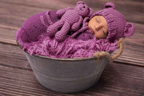 Neonata avvolta in una stoffa, con cappellino con orecchie da orsacchiotta, pupazzetto orso accanto a lei, in un secchio di metallo con copertina pelosa. tutto in sfumature di rosa e lilla. Sfondo tavolo di legno